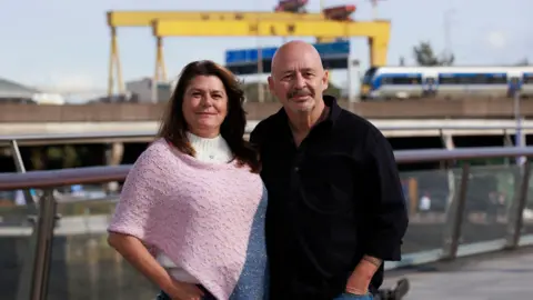 PA Media A woman in a pink top and a man in a black shirt. Yellow Harland & Wolff cranes are in the background.