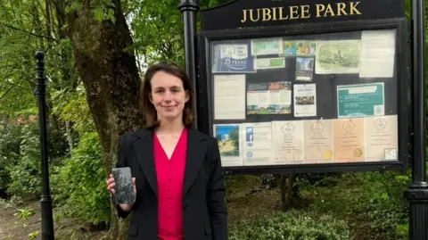 LDRS Elsie Blundell, wearing a black blazer over a red top, holds up her mobile phone. She is seen standing in front of a large noticeboad and sign for Jubilee Park