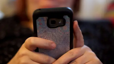 PA Media Generic close-up image of a woman holding her mobile phone in front of her. It has a shiny silver cover.