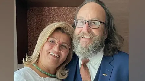 Supplied Lili and Dave Myers. Lili is leaning her head on Dave's shoulder an both are smiling. Lili has blonde, shoulder-length hair and a pale jade-green bead necklace. Dave is wearing a royal blue suit with salmon pink tie, with gold tie pin. He has shoulder length dark grey hair and his trademark full beard and moustache.