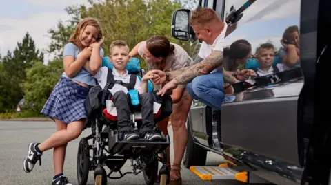 Family A family of four are stood around a mini-van. On the left is a girl who is holding hands with a boy who is in a wheelchair. The boy's parents are to the right and looking towards him