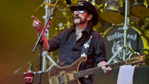 PA Media Lemmy wearing a black cowboy hat, a black shirt and holding a bass guitar, points with his right hand while he performs on stage.