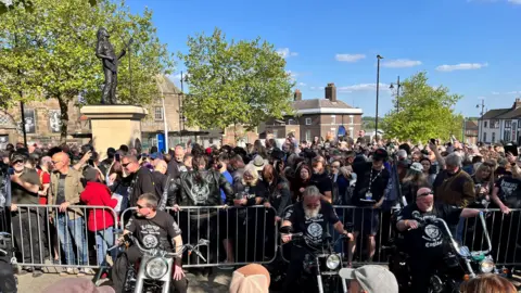 Three men on bikes in front of a railing, behind which is a crowd of people next to the new statue of Lemmy.