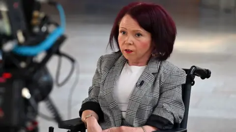 Getty Images A woman with red hair in the Scottish Parliament. She is sitting in a black wheelchair and is wearing a chequered grey suit jacket and white top.
