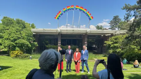 Two men and two women stand on the lawn outside a one-story exhibition centre with a rainbow spelling out "WE ARE POEMS". 