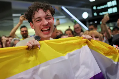 Reuters Last year's winner Nemo holding a non-binary flag and smiling to the camera at Zurich airport. 