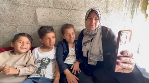 Three children look at a phone next to their grandmother, sitting against a wall