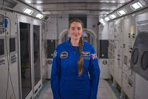 Kevin Church/BBC Picture of astronaut Rosemary Coogan in the replica of the International Space station at Johnson Space Centre. She is in the middle of the shot and has brown hair with a long plait that hangs down over her left shoulder. She is wearing a blue jumpsuit with a Union Jack on her left arm her name, and the European Space Agency logo on the front and the badge for her astronaut group " the hoppers" on her right arm.