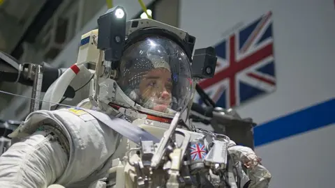Kevin Church/BBC Close up picture of Rosemary Coogan in her full space suit, with her helmet on looking straight ahead just before she begins her dive. A light is attached to the helmet on the right hand side. A small Union Jack is visible on the front of her spacesuit and there is a large Union Jack flag that is partially visible on the wall over her left shoulder.