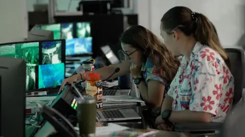 Kevin Church/BBC Trainers in the control room are leaning in to a microphone and communicating with the astronauts. There is a bank of screens in front of them. The woman on the left is wearing a blue flowery shirt and has brown hair and glasses. The woman on the right is wearing a white shirt with red stylised flowers. The dive is taking place on " wear a Hawaiian shirt to work day."