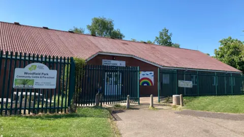 Outside of the Woodfield Park community centre and pre-school, it is a single-storey building with green gates outside.