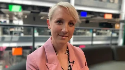 Emma Pinchbeck, chief executive of the Climate Change Committee sat in BBC Wales' headquarters in Cardiff, wearing a pink suit. She has cropped, blonde hair.