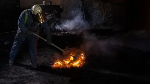 Getty Images A worker dressed in a blue overall and white helmet raking hot coal in blast furnace number four at Tata's Port Talbot steelworks.