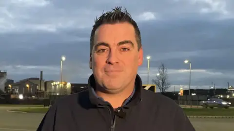 Shaun Spencer, a former worker at Tata's Port Talbot steelworks - which is visible behind him. He has short dark hair and is wearing a black fleece.