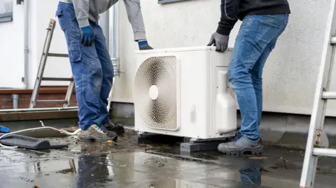 Getty Images Two technicians installing an outdoor heat pump unit on a wet surface. They are wearing jeans and working boots and are holding the white box with a circular vent on the side. You can only see their torso and legs in the image.