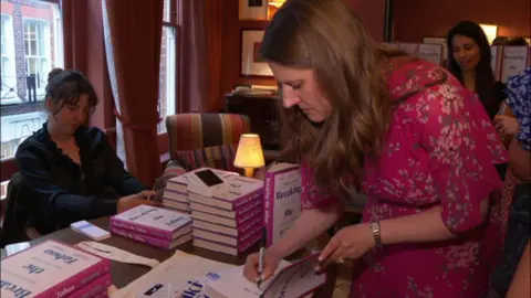 A woman with long dark hair leans over a desk to sign a book. She holds a pen in her right hand and wears a magenta dress with a flowery pattern on it. On her left wrist is a wristwatch. She signs the book which is among several piles of other books.