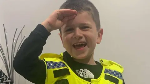 Family handout Archie give a salute with his right hand held up to his forehead while wearing a black top and toy police vest. The vest is bright yellow and had blue and white checked badges across the shoulders