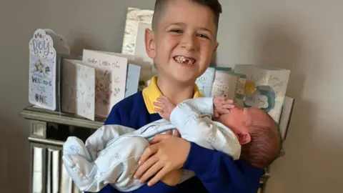 Family handout Archie smiles at the camera while holding his baby brother Finley in his arms. Archie has short dark hair and his big grin reveals he is missing his front teeth. He is wearing a school uniform of a yellow polo shirt and blue jumper. Finley has dark hair and is wearing a blue onesie. Behind them is a shelf covered in cards celebrating Finley's birth.