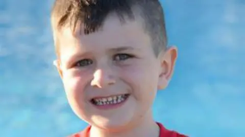 Family handout Archie smiles at the camera. He has short dark hair and is wearing a red t-shirt, with the blue waters of a swimming pool behind him.