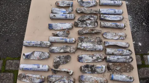 Northumbria Police 34 butane cannisters laid out on a board of wood. They are in varying conditions, some just reduced to ragged metal, others still showing the white and blue branding and distinctive cylindrical shape