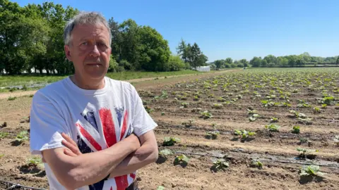 Malcolm Prior/BBC A man stands with his arms crossed wearing a white t-shirt with a union jack on it. Behind him is a dry-looking field with some small plants in rows in the soil.