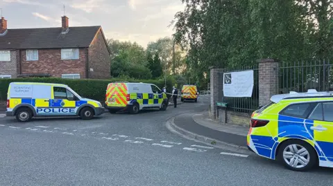 Merseyside Police officers and vehicles inside a cordon following an incident around the Tobruck Road area in Huyton on a sunny evening.