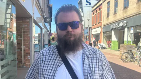 Harvey Bell/BBC Callum Kempton in a chequered overshirt and white T-shirt standing on Ipswich
High Street. He has reflective sunglasses and a brown bushy beard.