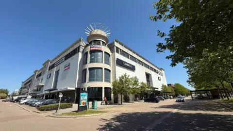 Laura Foster/BBC The exterior of the former Debenhams in Milton Keynes, with a large turret-shaped building in the middle surrounded by two large rectangular buildings. The sides of the buildings say Sports Direct and Flannels. In the foreground is a car park.