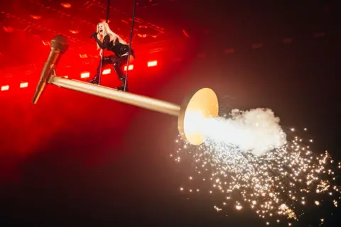 Sarah Louise Bennett / EBU Erika Vikman stands on a giant gold prop microphone, as fireworks explode around her