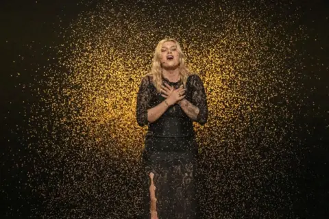 EPA-EFE/Shutterstock France's Eurovision entry Louane crosses her hands across her chest as she sings with her eyes closed, wearing a black dress and surrounded by a gold confetti-like effect, with a black backdrop.