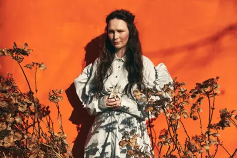 PA Media Swiss Eurovision entry Zoë Më stands in a posed mage in a white dress with an orange background, surrounded by flowers.
