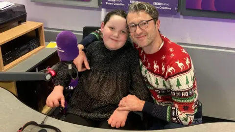 Justin Dealey/BBC Betsy is sitting in a chair in a radio studio with her father kneeling beside her. They are both smiling at the camera. Betsy is wearing a sparkly black top and her father is wearing spectacles and a Christmas jumper with reindeer and Christmas trees on it.