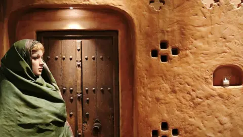 An installation that depicts the living conditions of Iranian Zoroastrians in Yazd shows a mannequin draped with a traditional Parsi shawl sitting outside a mud house, with a wooden door behind her. 