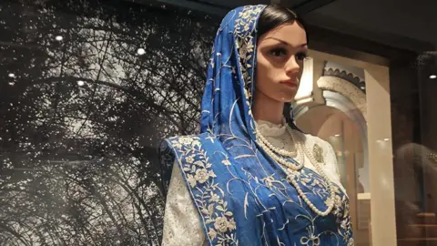 A mannequin draped in a blue traditional Parsi Ghara saree with floral motifs and pearl necklaces stands in a museum backdrop. 