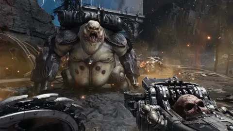 Bethesda Screenshot shows a first-person view of Doom: The Dark Ages' game play. A massive creature with frog-like trunk and turtle-esque head roars at the player character, exposing giant fangs. Two giant cannons take the place of its arms and it wears a metallic backpack - presumably feeding ammo to the weapons. The player holds a shield lined with saw blades on one side and an elaborate gun bedecked with a skull on the other.