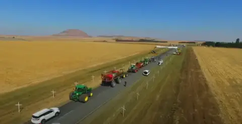 X Rows of white crosses can be seen on either side of a rural road in South Africa. Tractors and cars drive down the middle of the track, with fields on either side .