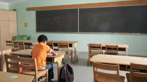 Getty Images A child is sitting at a desk alone. There are five desks in the room. There is a blackboard on the front wall, the board is divided in two. The wall of the room is painted green. The child is wearing an orange top with jeans. Their backpack is black and is sitting beside them. There is also a large wooden cupboard in the background. 