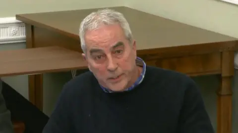 PA Media Pat Sheehan is wearing a blue shirt and navy jumper. He has grey hair. In the background there is two wooden tables. The walls are painted green and have a white feature running along them. 