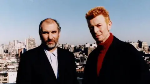 Changes: Bowie At Fifty - Picture shows Alan Yentob (left) and David Bowie. Yentob. The pair, both wearing black suits, are pictured with the London skyline behind them.
