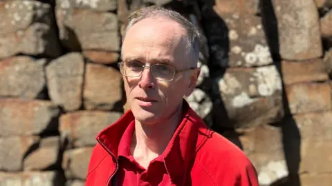 Dr Henry is looking at the camera and is wearing thin framed glasses. He has thin grey hair and is wearing a bright red fleece and polo top. 