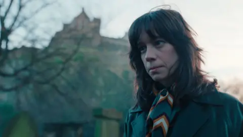 Netflix Close up of a woman staring at the ground with a serious expression on her face with a blurred Edinburgh castle visible in the background 
