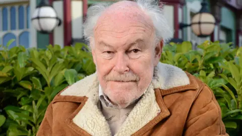 BBC Actor Timothy West looks at the camera. He has a grey beard and grey hair. He is wearing a tan coloured coat with a sheepskin collar.