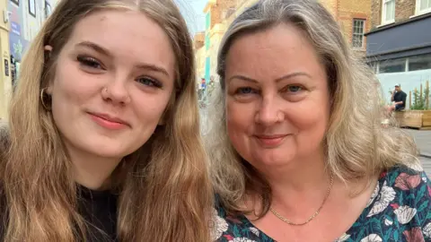 Amanda Two women smile at the camera. On the left a teenage girl with blonde hair and on the right a middle aged lady with a floral top smiles at the camera. She also has blonde hair.