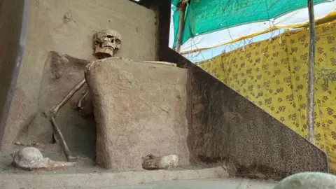Kushal Batunge/BBC A photo of the seated skeleton inside a make-shift shed which shows its right arm resting on its lap and its left arm suspended in the air, as if resting on a stick