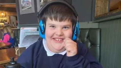 Handout Thomas, 12, who has black hair and is wearing blue ear defenders and a blue jumper, smiles at the camera while sitting on a table at a restaurant
