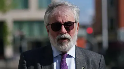 PA Media Gerry Adams with grey hair and sungalsses. He is wearing a grey suit, blue shirt and purple tie. He is outside.
