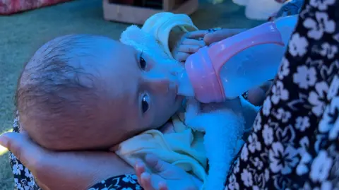 A malnourished-looking baby's head is held in the palm of a hand as she drinks from a milk bottle. 