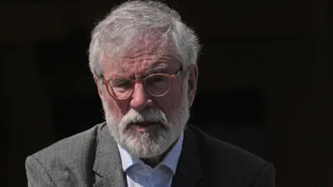 Brian Lawless/PA Wire Gerry Adams pictured outside the High Court in Dublin last week wearing a grey suit jacket, a light coloured shirt with the top button undone and a pair of circular brown glasses. Mr Adams has white hair and a beard.