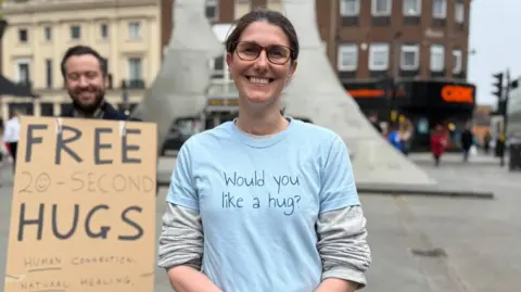 Danny Fullbrook/BBC A smiling woman wearing glasses and a blue t-shirt with the words "Would you like a hug?" stands in the street in front of a man, who is also smiling, holding a sign that reads "FREE 20-SECOND HUGS".