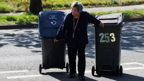 Reuters A woman pulls her wheeled bins to a Mobile Household Waste Centre where residents can dispose of their rubbish. She is wearing a dark top and trousers and has a lanyard around her neck. She is pulling two bins and stooping slightly. The sun is out and there is grass behind her and a tree trunk.